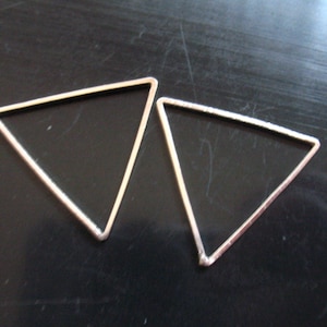 Op de afbeelding: Twee zilverkleurige, open driehoekvormen. De driehoeken zijn identiek in grootte en vorm, met een strak, geometrisch ontwerp. De randen zijn glad en het algehele uiterlijk is minimalistisch. Waarschijnlijk sieradenonderdelen of decoratieve elementen.