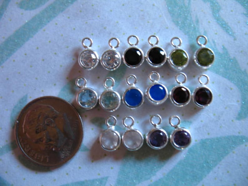 May include: Assortment of 15 silver charm pendants with various colored gemstones. The charms feature a loop at the top for attaching to a necklace or bracelet. The gemstones include clear, blue, green, purple, and black.