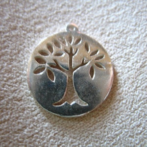 May include: Silver pendant in the shape of a circle with a tree design cut out. The tree has a thick trunk and branches with leaves.