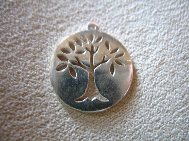 May include: Silver pendant in the shape of a circle with a tree design cut out. The tree has a thick trunk and branches with leaves.