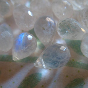 May include: A collection of iridescent moonstone teardrop beads. The beads are translucent and have a milky white colour with a blue sheen.