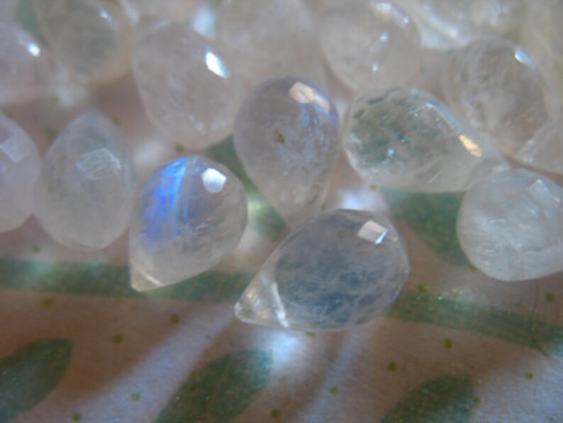 May include: A collection of iridescent moonstone teardrop beads. The beads are translucent and have a milky white colour with a blue sheen.