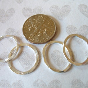 以下が含まれることがあります： わずかに異なる形をした銀色のハンマーで叩かれた金属製のリングが4つ、ハート模様の白い背景の上に並んでいます。サイズ比較のために、アメリカの25セント硬貨が添えられています。