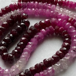 May include: A strand of faceted gemstone beads in shades of pink, red, and purple. The beads are arranged in a graduated color pattern, with the darkest beads at the bottom and the lightest beads at the top.