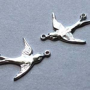 Może przedstawiać: Dwa srebrne metalowe wisiorki w kształcie jaskółki z kółkami do zawieszenia. Ptaki są zwrócone w przeciwne strony.