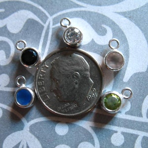 May include: Six silver charm pendants with different colored gemstones. The charms are arranged around a US dime coin. The gemstones are black, blue, clear, green, and white.
