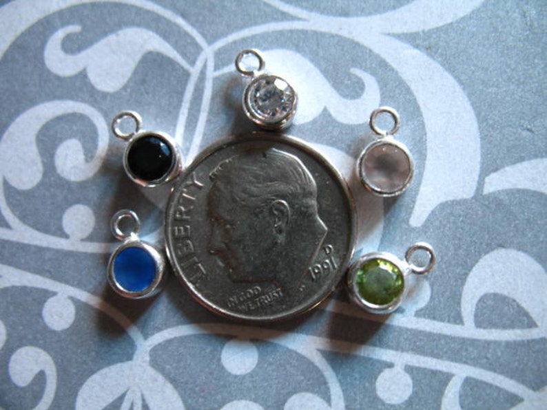 May include: Six silver charm pendants with different colored gemstones. The charms are arranged around a US dime coin. The gemstones are black, blue, clear, green, and white.