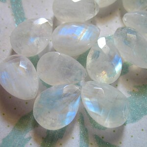 May include: A collection of iridescent moonstone gemstone beads in various shapes and sizes. The beads are a milky white colour with a rainbow sheen.