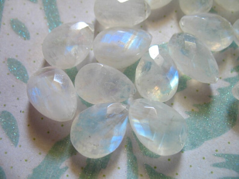 May include: A collection of iridescent moonstone gemstone beads in various shapes and sizes. The beads are a milky white colour with a rainbow sheen.