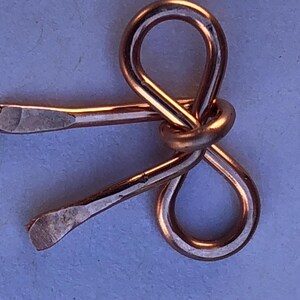 May include: A copper-colored metal bow tie, crafted from a single piece of wire, with a smooth, polished finish.