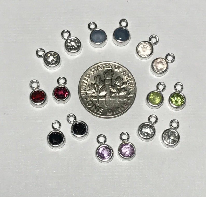 May include: A collection of 14 small silver charms with different colored gemstones. The charms are arranged in a circle around a US dime.