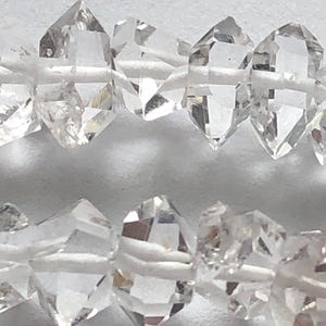 May include: A strand of clear quartz crystal beads, each bead is faceted and has a pointed shape. The beads are strung on a white cord.