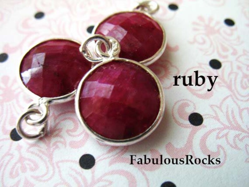 May include: Three ruby red faceted gemstone beads with silver connectors. The beads are connected to form a pendant or charm. The word "ruby" is written in white text on a pink and white background.