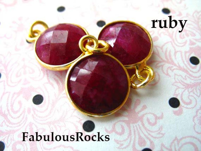 May include: Three ruby red gemstone beads with gold accents. The beads are faceted and have a shiny finish. The word "ruby" is written in black text above the beads.