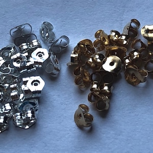 May include: A collection of earring backs in silver and gold tones. The silver backs are clustered on the left, while the gold backs are on the right. The earring backs are in various shapes, including butterfly and disc styles.