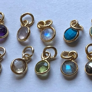 May include: A collection of ten gold-colored gemstone charms. The charms feature various colored gemstones, including amethyst, rose quartz, moonstone, turquoise, and peridot. Each charm has a small loop for attaching to a necklace or bracelet.