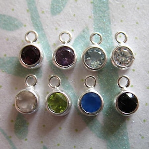 May include: Eight silver charms with different colored gemstones. The charms are round and have a loop at the top for attaching to a necklace or bracelet. The gemstones are amethyst, aquamarine, clear, green, black, white, blue, and red.