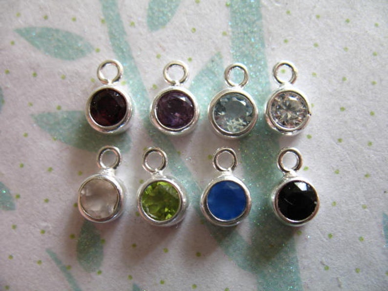 May include: Eight silver charms with different colored gemstones. The charms are round and have a loop at the top for attaching to a necklace or bracelet. The gemstones are amethyst, aquamarine, clear, green, black, white, blue, and red.