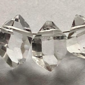Puede incluir: Primer plano de cuatro cuentas de cristal de cuarzo transparente enhebradas en un alambre de plata. Las cuentas son facetadas y tienen una forma de diamante.