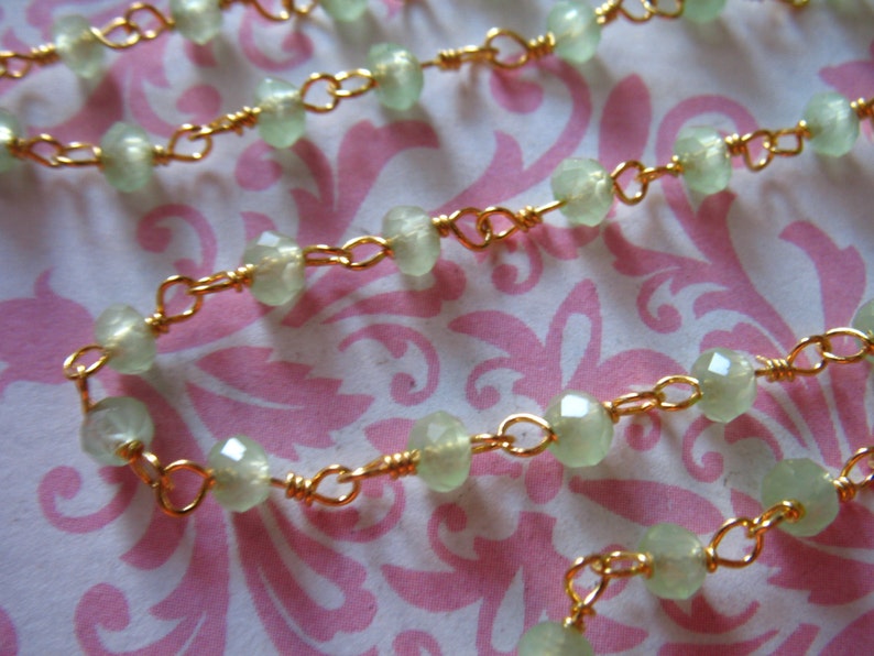May include: A delicate gold chain necklace with light green faceted beads. The necklace is on a pink and white patterned background.