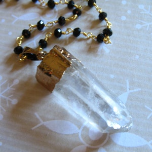 May include: A gold chain necklace with black beads and a clear quartz crystal pendant. The pendant is wrapped in gold metal.