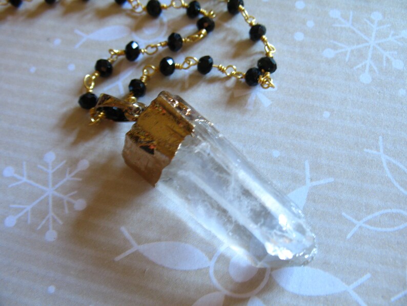 May include: A gold chain necklace with black beads and a clear quartz crystal pendant. The pendant is wrapped in gold metal.