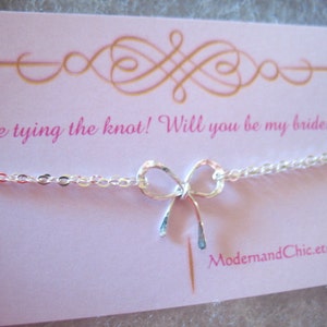 May include: A white card with gold lettering that says "We are tying the knot! Will you be my bridesmaid?" with a silver chain and bow charm. The card has the website "ModernandChic.etsy.com" printed on the bottom.