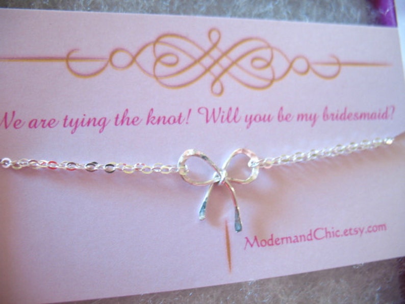 May include: A white card with gold lettering that says "We are tying the knot! Will you be my bridesmaid?" with a silver chain and bow charm. The card has the website "ModernandChic.etsy.com" printed on the bottom.