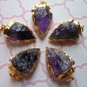 Puede incluir: Cinco colgantes de punta de flecha de amatista con bordes chapados en oro. Las puntas de flecha son de diferentes tamaños y formas, pero todas son de color morado oscuro.