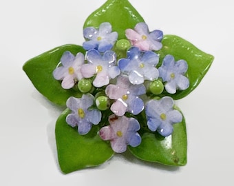 Peut inclure: Une broche en porcelaine représentant un bouquet de fleurs bleues et roses de myosotis avec des feuilles vertes.