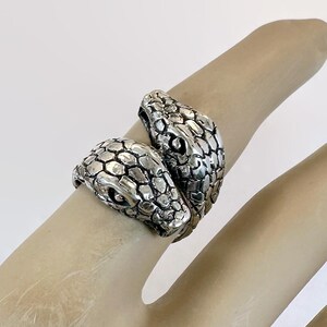 May include: A silver ring featuring two snake heads facing each other. The ring has a detailed, textured design, with scales and eye details. The ring is worn on a finger, showcasing the jewelry's design.
