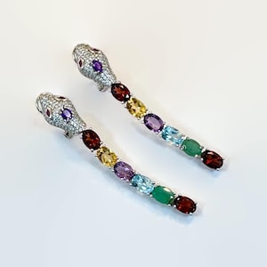 May include: A pair of silver-toned snake earrings. The snake heads are encrusted with small, clear stones and feature red and purple accents. The bodies are composed of a series of colorful, oval-shaped gemstones in red, yellow, purple, blue, green, and garnet.