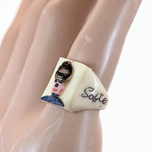 Könnte beinhalten: Ein cremefarbener Ring mit einer handbemalten Illustration einer Figur mit dunklem Haar und blauem Kragen auf der Vorderseite. Die Seite des Rings ist mit dem in Schreibschrift verfassten Wort "Softie" in schwarzer Tinte auf glatter Oberfläche versehen.