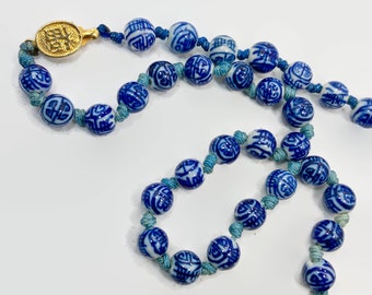May include: A blue and white porcelain bead necklace with a gold clasp. The beads are round and have Chinese characters on them. The necklace is strung on a blue cord.