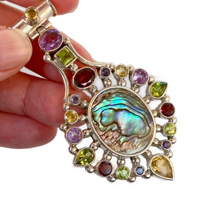 Può includere: Un ciondolo d'argento con una grande conchiglia di abalone iridescente circondata da un alone di pietre preziose colorate, tra cui ametista, peridoto, citrino e granato.