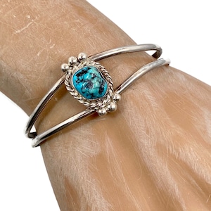 Puede incluir: Un brazalete de plata con una piedra turquesa engastada en un bisel de plata. El brazalete tiene un diseño de doble banda y está adornado con pequeñas cuentas de plata.