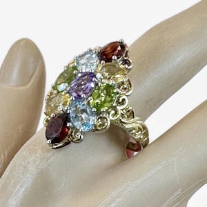 Puede incluir: Un anillo dorado con un grupo de piedras preciosas multicolores. El anillo presenta piedras de corte ovalado en tonos rojos, azules, verdes, amarillos y morados. El anillo tiene un diseño ornamental y ondulado.