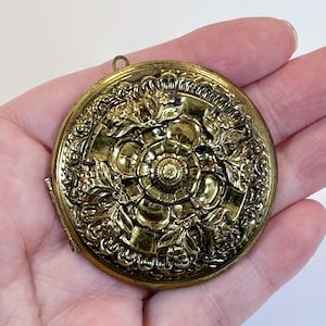 May include: A round, antique gold-colored locket with intricate floral and leaf detailing. The locket is open, revealing a small interior space. The design features a central flower surrounded by ornate embellishments. A small loop is at the top for a chain.