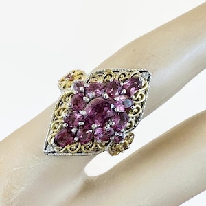 Könnte beinhalten: Ein silberner und goldener Ring mit einem Cluster aus oval geschliffenen, tiefvioletten Edelsteinen. Der Ring hat ein verziertes Filigran-Design um die Edelsteine und an den Schultern des Rings. Der Ring ist auf einem hellen Hintergrund gesetzt.