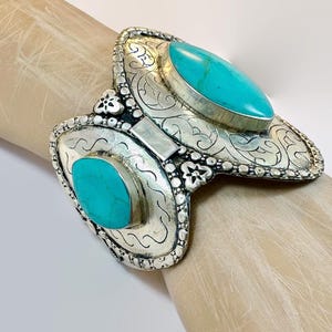 May include: Silver-toned cuff bracelet with two turquoise stones. The bracelet features intricate scrollwork and decorative details. The turquoise stones are oval and rectangular in shape. The bracelet is worn on a wrist.