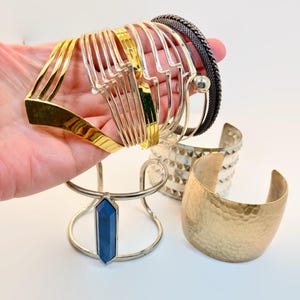 May include: A collection of various cuff bracelets including a gold-toned multi-wire cuff, a dark metallic mesh cuff, a silver-toned cuff with a central blue hexagonal stone, a silver-toned cuff with pyramid studs, and a wide hammered gold-toned cuff.