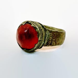 May include: Antique ring with a large, round, red gemstone set in a tarnished metal band. The band features decorative etched details around the gemstone and on the side. The ring's design suggests a vintage or historical style.