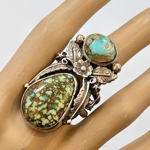 May include: A silver ring featuring two large turquoise stones with brown matrix patterns. The ring has a floral and leaf design, with a detailed setting. The turquoise stones are oval and teardrop shaped.