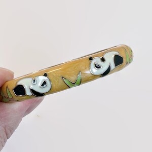 May include: A yellow enamel bangle bracelet featuring panda bear designs. The bracelet has a light yellow background with black, white, and green panda illustrations and bamboo accents. The bracelet is held by a hand.