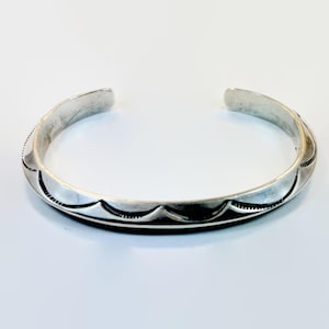 Puede incluir: Una pulsera de plata con un diseño abierto. La pulsera presenta un patrón repetitivo de líneas curvas y pequeños detalles grabados a lo largo de los bordes. El metal tiene un aspecto ligeramente envejecido.
