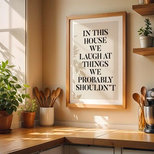 Puede incluir: Una cocina con un cuadro enmarcado con el texto "IN THIS HOUSE WE LAUGH AT THINGS WE PROBABLY SHOULDN'T". La cocina cuenta con estantes de madera con plantas en macetas, utensilios de madera y una batidora de pie negra.