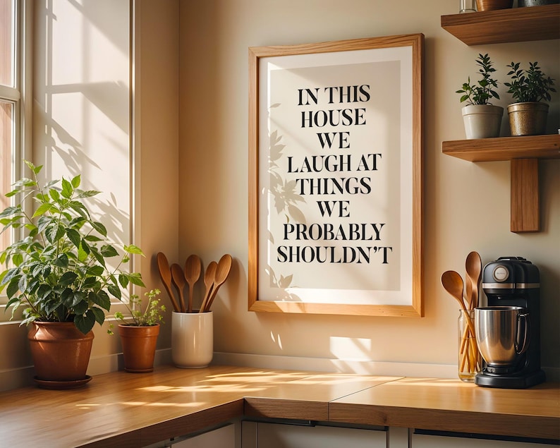 Puede incluir: Una cocina con un cuadro enmarcado con el texto "IN THIS HOUSE WE LAUGH AT THINGS WE PROBABLY SHOULDN'T". La cocina cuenta con estantes de madera con plantas en macetas, utensilios de madera y una batidora de pie negra.