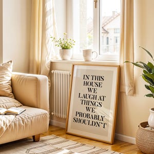 Puede incluir: Un cuadro enmarcado con el texto "IN THIS HOUSE WE LAUGH AT THINGS WE PROBABLY SHOULDN'T" se apoya contra una pared. Un sof&aacute; beige con un libro, una planta en maceta y una cesta de mimbre est&aacute;n en la habitaci&oacute;n. Una ventana con cortinas est&aacute; al fondo.