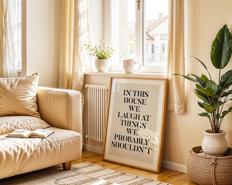Puede incluir: Un cuadro enmarcado con el texto "IN THIS HOUSE WE LAUGH AT THINGS WE PROBABLY SHOULDN'T" se apoya contra una pared. Un sof&aacute; beige con un libro, una planta en maceta y una cesta de mimbre est&aacute;n en la habitaci&oacute;n. Una ventana con cortinas est&aacute; al fondo.