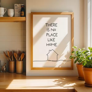 May include: A wooden framed print with the text "THERE IS NO PLACE LIKE HOME" and a house illustration. The frame is on a wooden countertop with potted plants and kitchen utensils. A stainless steel kettle is on the left.